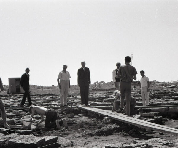 Den svartvita bilen visar några silhuetter av människor vid en arkeologisk utgrävning i ett kargt landskap.