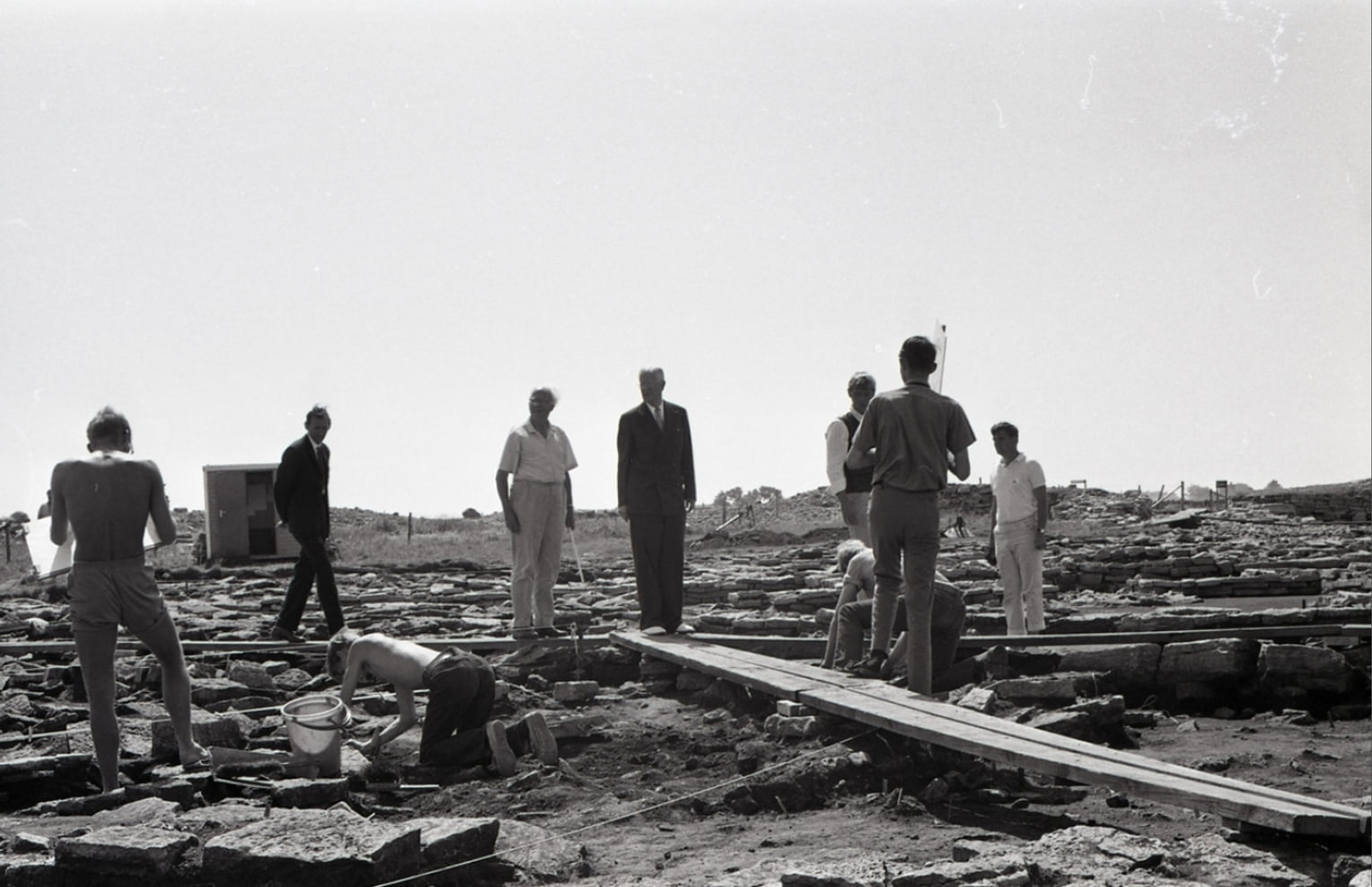 Den svartvita bilen visar några silhuetter av människor vid en arkeologisk utgrävning i ett kargt landskap.