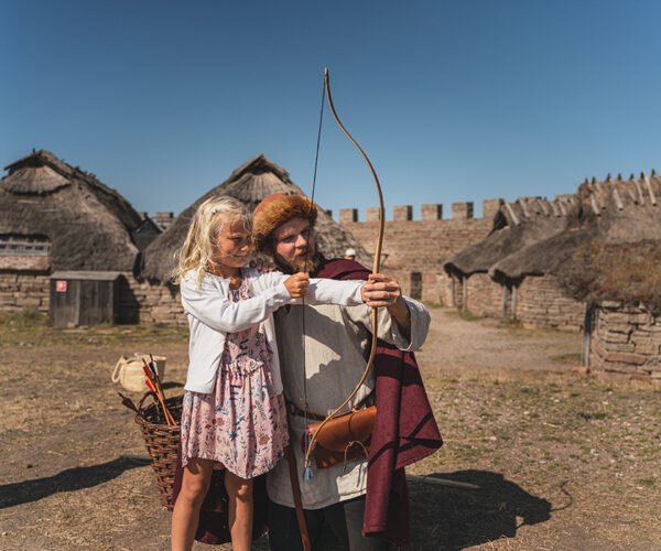 Ett barn skjuter pilbåge med hjälp av en guide i medeltida kläder,