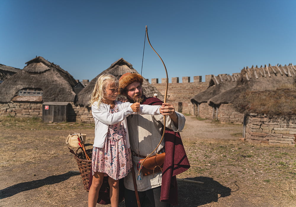 Ett barn skjuter pilbåge med hjälp av en guide i medeltida kläder,