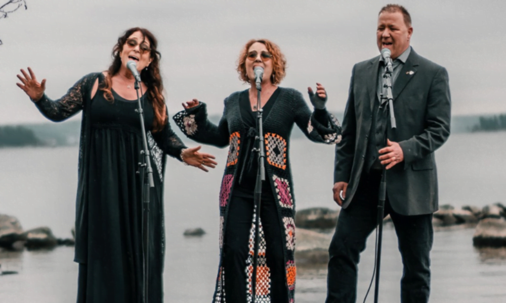 Tre personer står vid mikrofoner och sjunger. I bakgrunden syns en sjö.