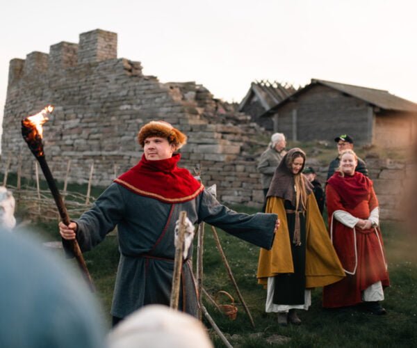En man i medeltida kläder håller i en fackla och berättar en historia för en grupp människor.